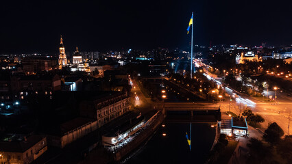 Flag of Ukraine on illuminated river embankment with water reflection at night. City aerial view above river Lopan near Skver Strilka and Cathedral in Kharkiv, Ukraine