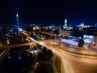 Flag of Ukraine on illuminated river embankment with reflection at night. City aerial view near Dormition Cathedral in Kharkiv, Ukraine with long exposure and background blur