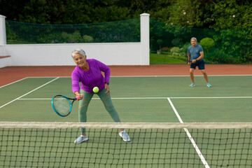Obraz premium Biracial active senior woman with senior man playing tennis in tennis court against trees