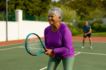 Smiling biracial senior woman with short hair playing tennis in court with senior man in background