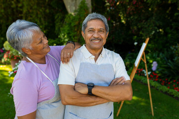 Smiling biracial senior woman with hand on husband's shoulder standing against plants in yard - Powered by Adobe