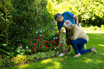 Biracial senior woman with senior man holding trowel and fork tools while gardening plants in yard