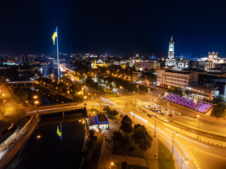 Fototapeta premium Flag of Ukraine on illuminated river embankment with reflection at night. City aerial view above river Lopan near Dormition Cathedral in Kharkiv, Ukraine with long exposure