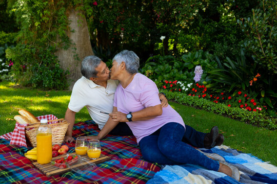 Biracial senior couple kissing while sitting with juice and food on blanket against plants in park