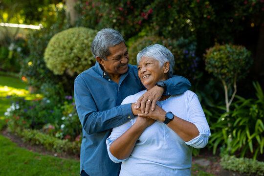 Smiling biracial senior couple embracing and holding hands while standing against plants in park