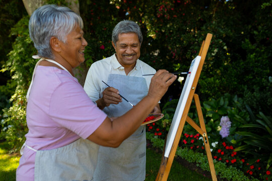 Cheerful biracial senior couple painting with watercolors on canvas against plants in yard