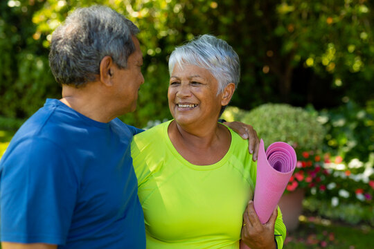 Smiling biracial senior woman holding yoga mat looking at husband while standing in yard