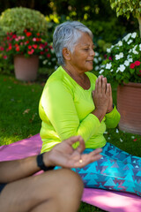 Biracial senior woman meditating with senior husband while sitting on mat against plants in yard