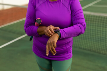 Midsection of biracial senior woman holding racket and checking time over wristwatch at tennis court - Powered by Adobe