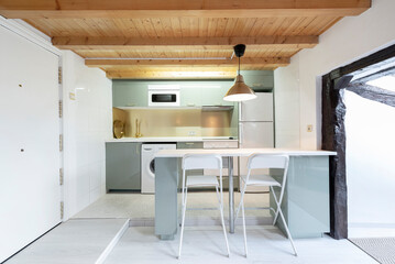 Kitchen with white and green cabinets in an apartment with sloped and beamed ceilings and wooden ceilings, matching island and tall white folding chairs
