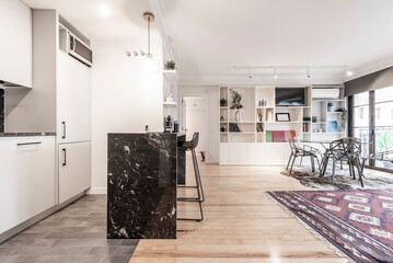 Integral kitchen with floor-to-ceiling white furniture, black marble top on the island with metal and black resin chairs and an open living room with a white bookcase