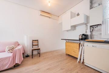 Apartment kitchen with wooden cabinets, black countertops, chestnut wood flooring, sofa with pink fabric sofa cover and air conditioner on the wall