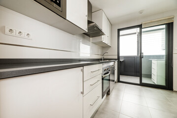 Apartment kitchen with white wooden furniture, black countertop, smooth stoneware floor and terrace...