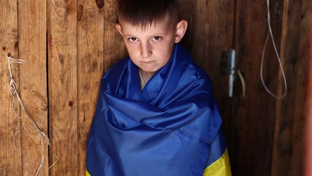 Ukrainian small child stands outdoors supporting homeland, little kid covered in national flag looking at camera. Protesting against Russian war in Ukraine, armed conflict, homeless, crisis, portrait.