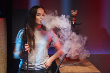 a young woman smokes a hookah exhaling a large white cloud of smoke. hookah next to her