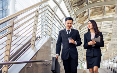 Two happy adult Asian businessman and woman or colleagues wearing formal suits, talking, walking together outdoor, going to work, holding cup of coffee with refreshment. Business, Lifestyle Concept.