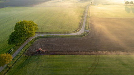 Aerial drone view of a tractor with cultivator plowing field in the early morning fog lit up the golden sunrise. Tractor disk harrow on ploughing a soil. Planting in farmland. Sowing seed on plowed