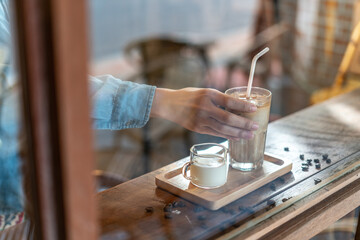 Fototapeta na wymiar A hand holding a tall glass of iced latte coffee with milk on a wooden bar over a cafe glass window reflex at a Cafe coffee shop. Cold brew refreshment summer drink with copy space. Selective focus
