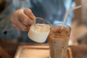 A hand pouring a glass of milk cream into iced latte coffee on a wooden bar over a cafe glass window reflex at a Cafe coffee shop. Cold brew refreshment summer drink with copy space. Selective focus