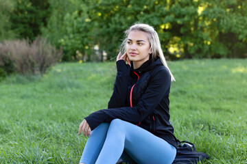 A beautiful girl sits on the grass after a workout. Cute model in sportswear in the park.