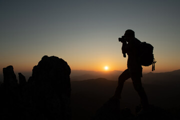 Silhouette of a travel photographer sitting on a rock and starting to shoot in the evening awaiting sunset at the peak of the national park. adventure experience concept