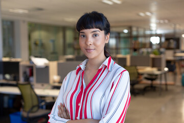 Fototapeta premium Portrait of confident young biracial businesswoman with arms crossed at modern workplace