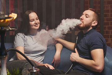 young man and woman are sitting on the sofa and smoking a hookah in a hookah lounge in a relaxed atmosphere. man exhales a cloud of white smoke