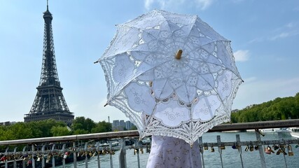 a girl with a white parasol stands against the backdrop of the Eiffel Tower with her back to us can...