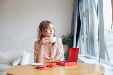 Attractive lady sitting at the table, drinking coffee or tea and eating chocolate at home