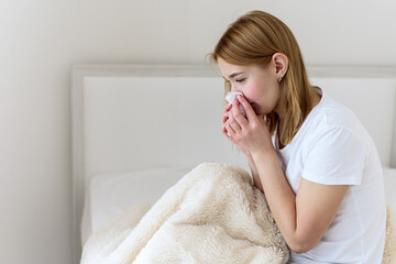 Sick young woman suffers from stuffy nose and sore throat. Upset sick Caucasian lady sitting in bed...