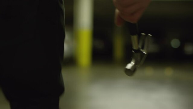 A man quickly aggressively walks through a car parking with a hammer in his hand. A tense scene of an upcoming crime. Thriller danger style. Character tracking effect. High quality closeup footage. 