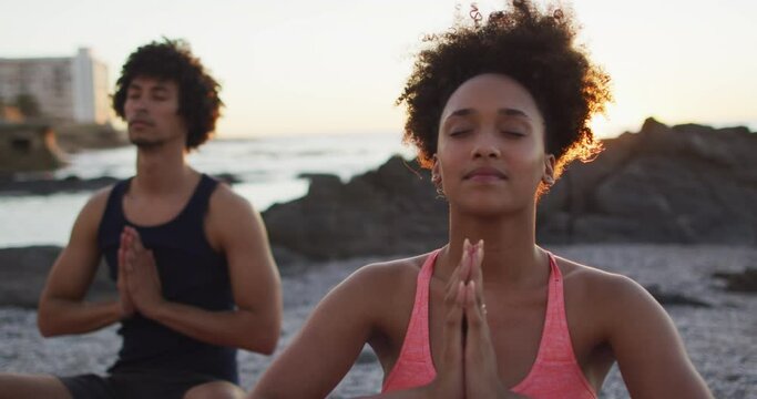 African american couple practicing yoga and meditating together on rocks near the sea during sunset