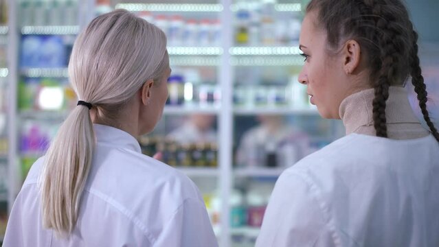 Back view intelligent serious women talking standing in front of shelves in drugstore. Professional Caucasian confident druggists chatting at workplace in pharmacy indoors