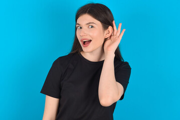 Fototapeta premium Oh my God! Funny astonished young caucasian woman wearing black T-shirt over blue background opening mouth widely looking aside, with hand near ear trying to listen to gossips.