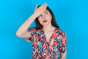 young caucasian woman wearing floral dress over blue background touching forehead, hears something surprising, glad receive good news, feels relieved. Almost got in trouble.