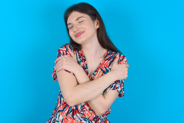 young caucasian woman wearing floral dress over blue background . Hugging oneself happy and positive, smiling confident. Self love and self care.