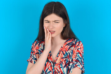 Tooth ache concept. young caucasian woman wearing floral dress over blue background feeling pain, holding his cheek with hand, suffering from bad toothache, looking at camera with painful expression
