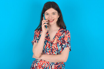 Portrait of a smiling young caucasian woman wearing floral dress over blue background talking on mobile phone. Business, confidence and communication concept.