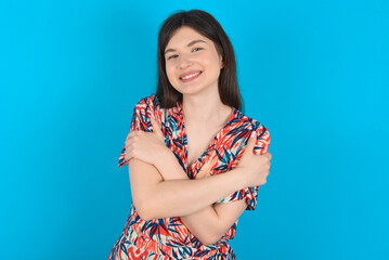 Charming pleased young caucasian woman wearing floral dress over blue background embraces own body, pleasantly feels comfortable poses. Tenderness and self esteem concept