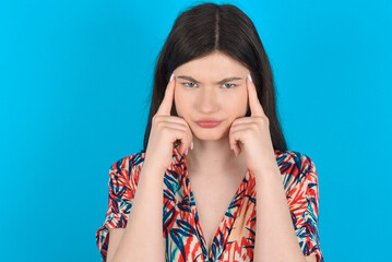 Serious concentrated young caucasian woman wearing floral dress over blue background keeps fingers on temples, tries to ease tension, gather with thoughts and remember important information for exam