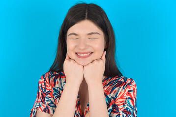 Cheerful young caucasian woman wearing floral dress over blue background has shy satisfied expression, smiles broadly, shows white teeth, People emotions