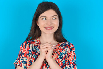 Happy young caucasian woman wearing floral dress over blue background anticipates something awesome happen, looks happily aside, keeps hands together near face, has glad expression.