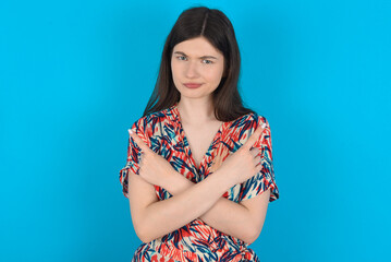Serious young caucasian woman wearing floral dress over blue background crosses hands and points at different sides hesitates between two items. Hard decision concept