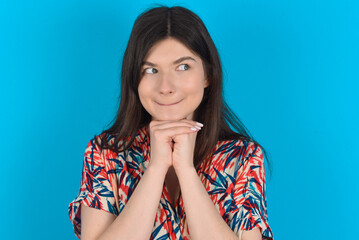 Curious young caucasian woman wearing floral dress over blue background keeps hands under chin bites lips and looks with interest aside.