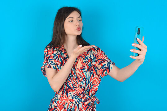 young caucasian woman wearing floral dress over blue background blows air kiss at camera of smartphone and takes selfie, sends mwah via online call.