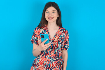 young caucasian woman wearing floral dress over blue background holds mobile phone in hands and rejoices positive news, uses modern cellular