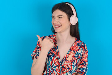 young caucasian woman wearing floral dress over blue background listens audio track via wireless headphones points thumb away advertises copy space