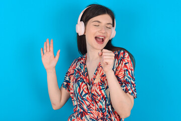 Fototapeta premium Happy young caucasian woman wearing floral dress over blue background sings favourite song keeps hand near mouth as if microphone wears wireless headphones, listens music