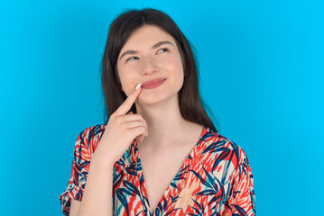 Lovely dreamy young caucasian woman wearing floral dress over blue background keeps finger near lips looks aside copy space.