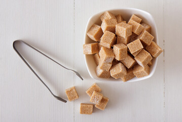 Cube-shaped cane sugar in wooden bowl on a white wooden table with silver sugar tongs. Flat lay, top down view, no people.